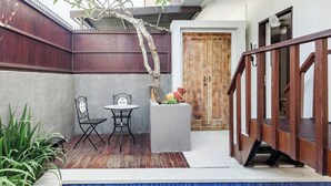 Un patio con una terraza de madera, dos sillas de metal, una mesita, un árbol ornamental y una puerta de madera.