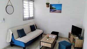 A modern living room with a sofa, coffee table, TV stand, and a painting of a beach scene. - T3 furnished, 2 air-conditioned bedrooms, terrace, located 5 minutes from southern beaches (Rivière-Pilote)