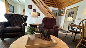 A cozy living room with a wooden ceiling, a black leather armchair, a red armchair, a wooden coffee table with decorative items, and a lamp. - Cozy 1-bedroom cabin with AC, WiFi & garage in charming Ironwood (Ironwood)