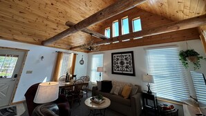 A cozy room with wooden ceiling beams, a ceiling fan, a chandelier, a dining table with chairs, a sofa, and a television. - Cozy 1-bedroom cabin with AC, WiFi & garage in charming Ironwood (Ironwood)