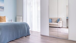 Ein Schlafzimmer mit Bett, Heizkörper, Fenster mit leichtem Vorhang und Kleiderschrank mit Spiegel.