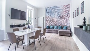 Ein modernes Wohnzimmer mit einem weißen Esstisch, grauen Stühlen, einem grauen Sofa und Kissen in Türkisgrün, sowie einem großen Wandbild mit abstrakten Blattmotiven.