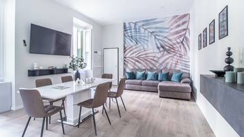 A modern living room with a white dining table, grey chairs, a grey sofa with teal cushions, and a large abstract leaf-patterned wall mural.