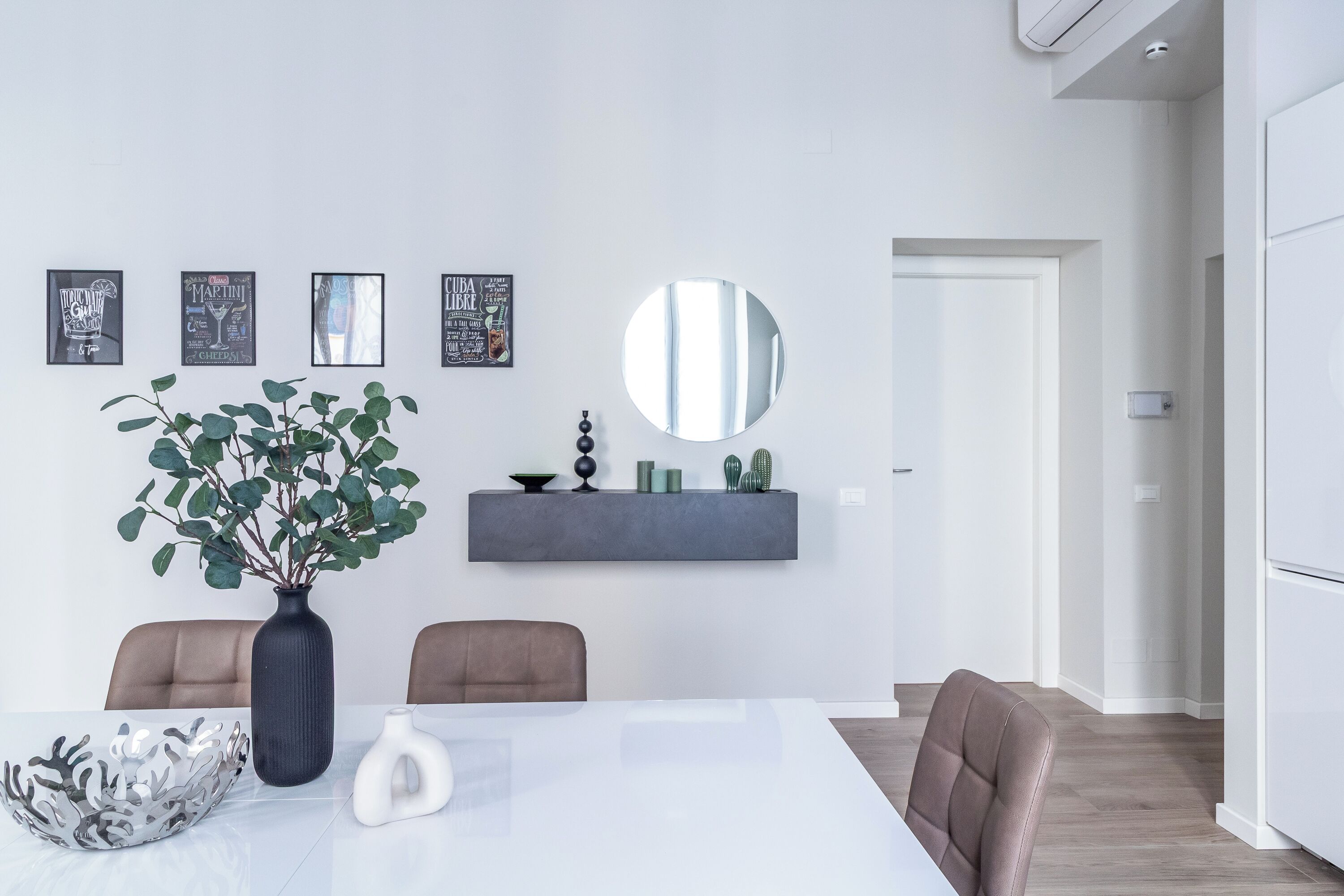 A modern dining area with a white table, two chairs, a vase with green foliage, and a wall-mounted shelf with decorative items.