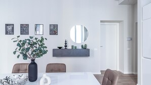 Ein moderner Essbereich mit einem weißen Tisch, zwei Stühlen, einer Vase mit Grünpflanzen und einem an der Wand befestigten Regal mit Dekorationen.