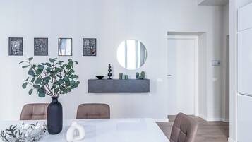 A modern dining area with a white table, two chairs, a vase with green foliage, and a wall-mounted shelf with decorative items.
