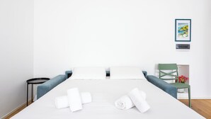 A neatly made bed with white linens, a small bedside table, a green chair, and a framed picture on the wall.