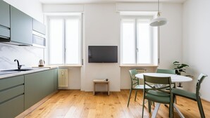 A modern kitchen with a built-in stove, a sink, and a small table with a kettle. There are two windows with white frames and a flat-screen TV mounted on the wall.
