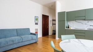 A modern living room with a blue sofa, a dining table with chairs, and a kitchen area with green cabinets and a marble countertop.
