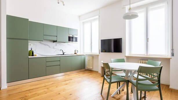 A modern kitchen with green cabinets, a white countertop, and a wooden floor. There is a dining area with a white table and green chairs.
