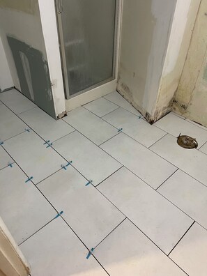 A room with white tiled flooring, a door with a glass panel, and visible wall damage.