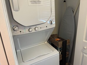 Un lave-linge à chargement frontal doté de panneaux de commande, un sèche-linge placé en dessous, et un panier en osier contenant un livre.