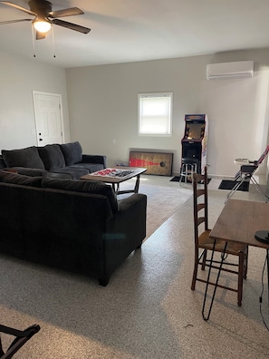 Un salon avec un canapé de couleur foncée, une table basse en bois, une petite table avec une chaise, un ventilateur de plafond et une fenêtre avec des stores.