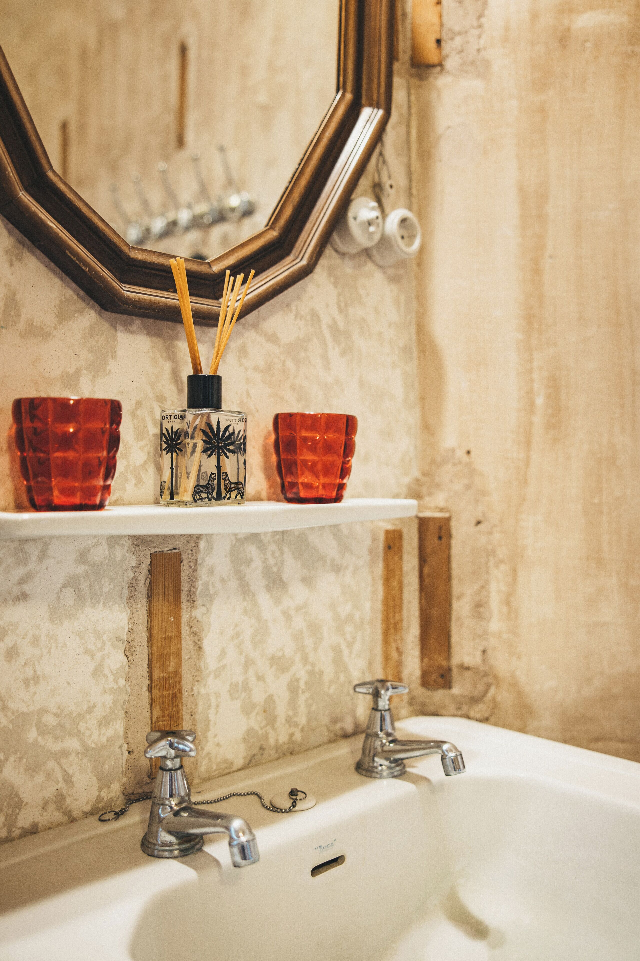 Un bagno con un lavandino bianco, due rubinetti, uno specchio e uno scaffale con due contenitori di vetro rossi e una confezione di deodorante per ambienti.