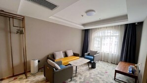 Ein Hotelzimmer mit einer Sofagarnitur, einem kleinen Tisch, einem Sessel, einem Mülleimer und einem Fenster mit Vorhängen.