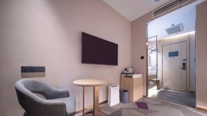 Ein Hotelzimmer mit einem grauen Sessel, einem kleinen runden Tisch, einem flachbildfernseher an der Wand, einem Mülleimer und einem durch eine geöffnete Tür einsehbaren Badezimmer.