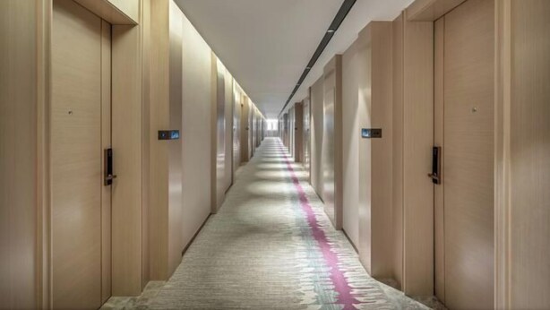 Ein langer, schmaler Hotelgang mit Holztüren an beiden Seiten, einem gemusterten Teppich und einer Deckenleuchte.