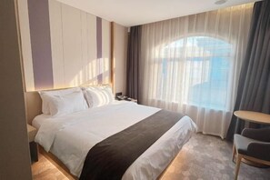 A hotel room with a bed, a nightstand, a chair, a small table, and a window with curtains. - Galaxy Hotel (Beijing Wukesong Wanshou Road Subway Station Branch) (Beijing)