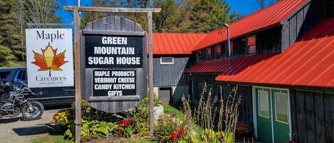 Ein rustikales Gebäude mit rotem Dach, mit der Aufschrift 'Green Mountain Sugar House', und einem Schild, das Ahornsirup-Produkte und Käse aus Vermont anbietet.