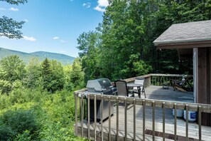 A wooden deck with a grill and outdoor furniture, surrounded by lush greenery and mountains. - Rustic cabin w/ mountain view & fireplace (Ludlow)