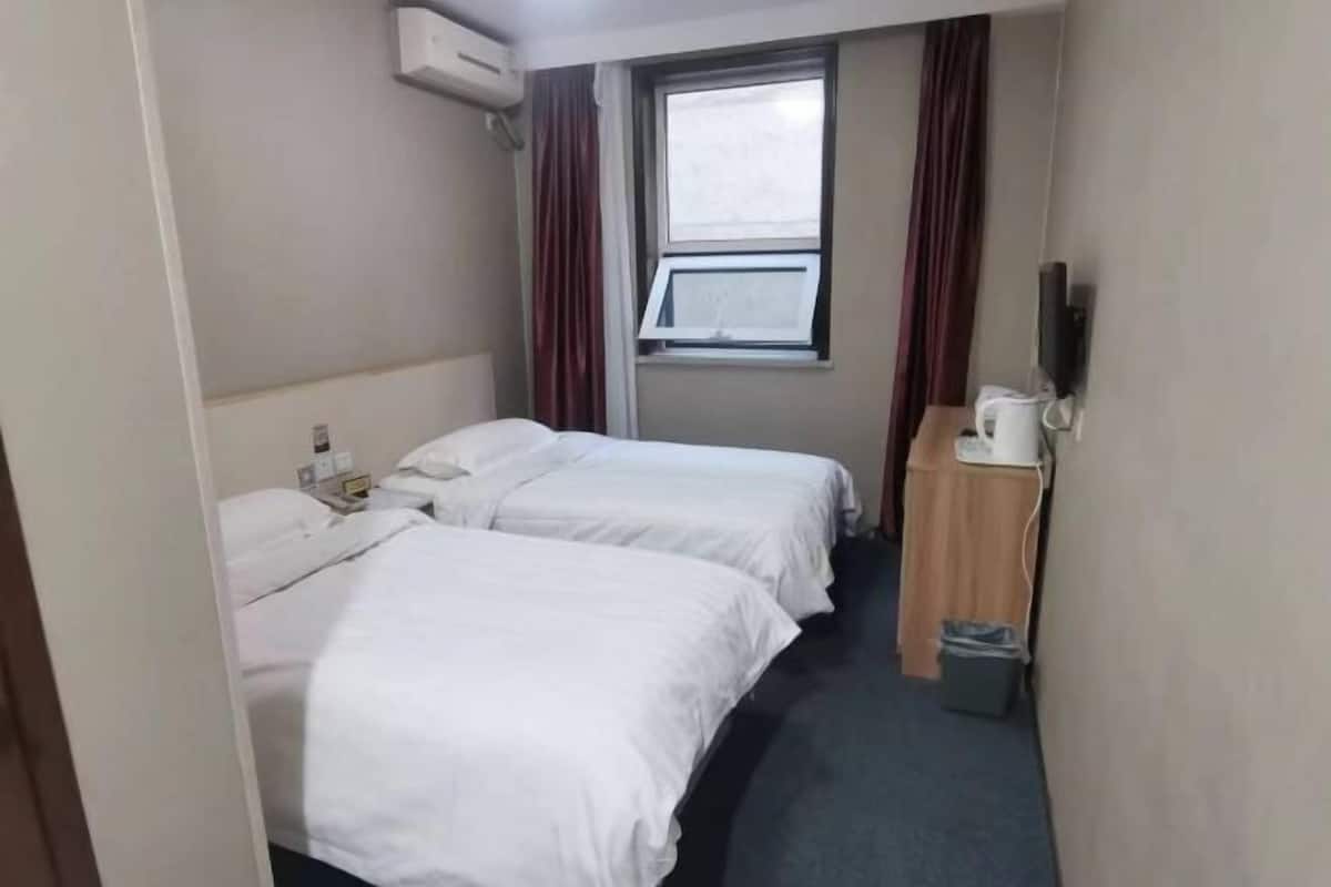 A small hotel room with two beds, a window with curtains, a desk with a telephone, and a trash bin.
