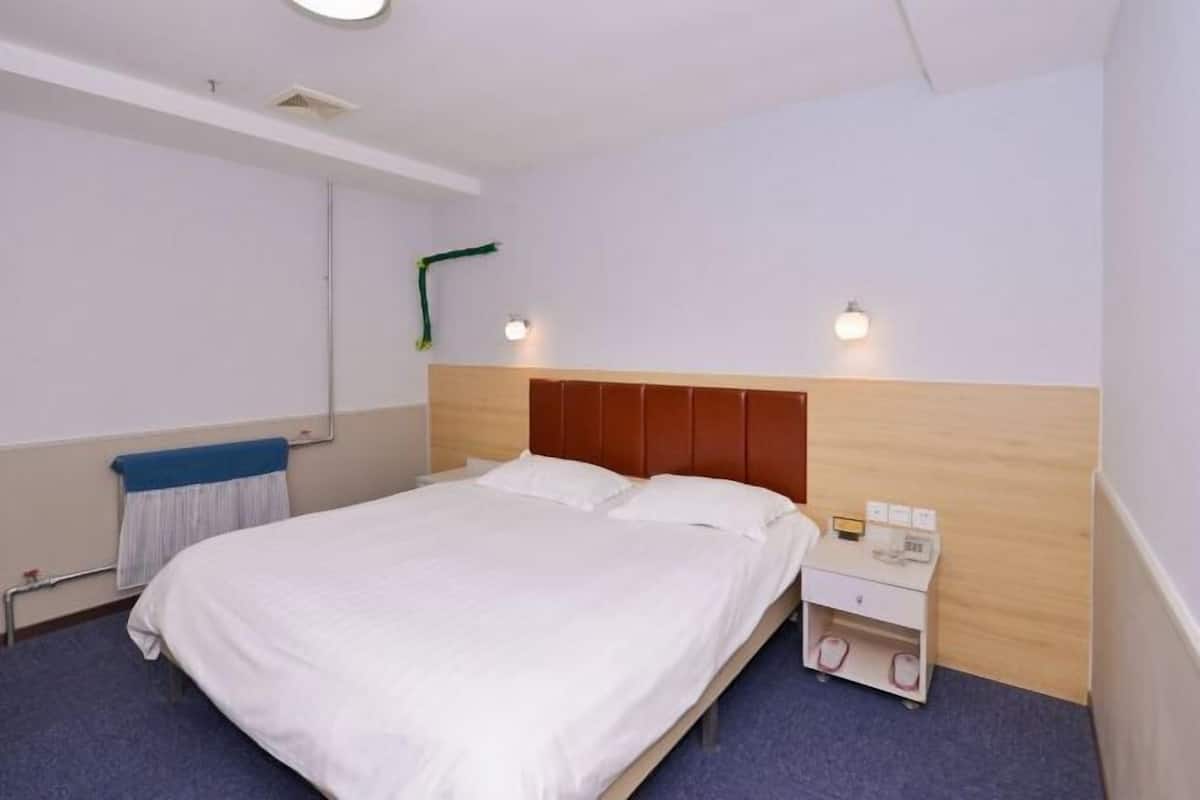 A hotel room with a single bed, a bedside table with a phone and a lamp, a wall-mounted light, and a towel rack.