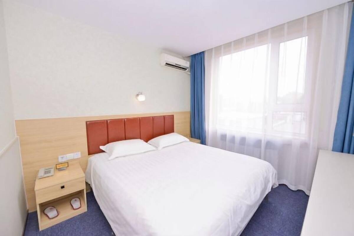 A hotel room with a bed, a nightstand, a shoe rack, a wall-mounted air conditioner, and a window with sheer curtains.