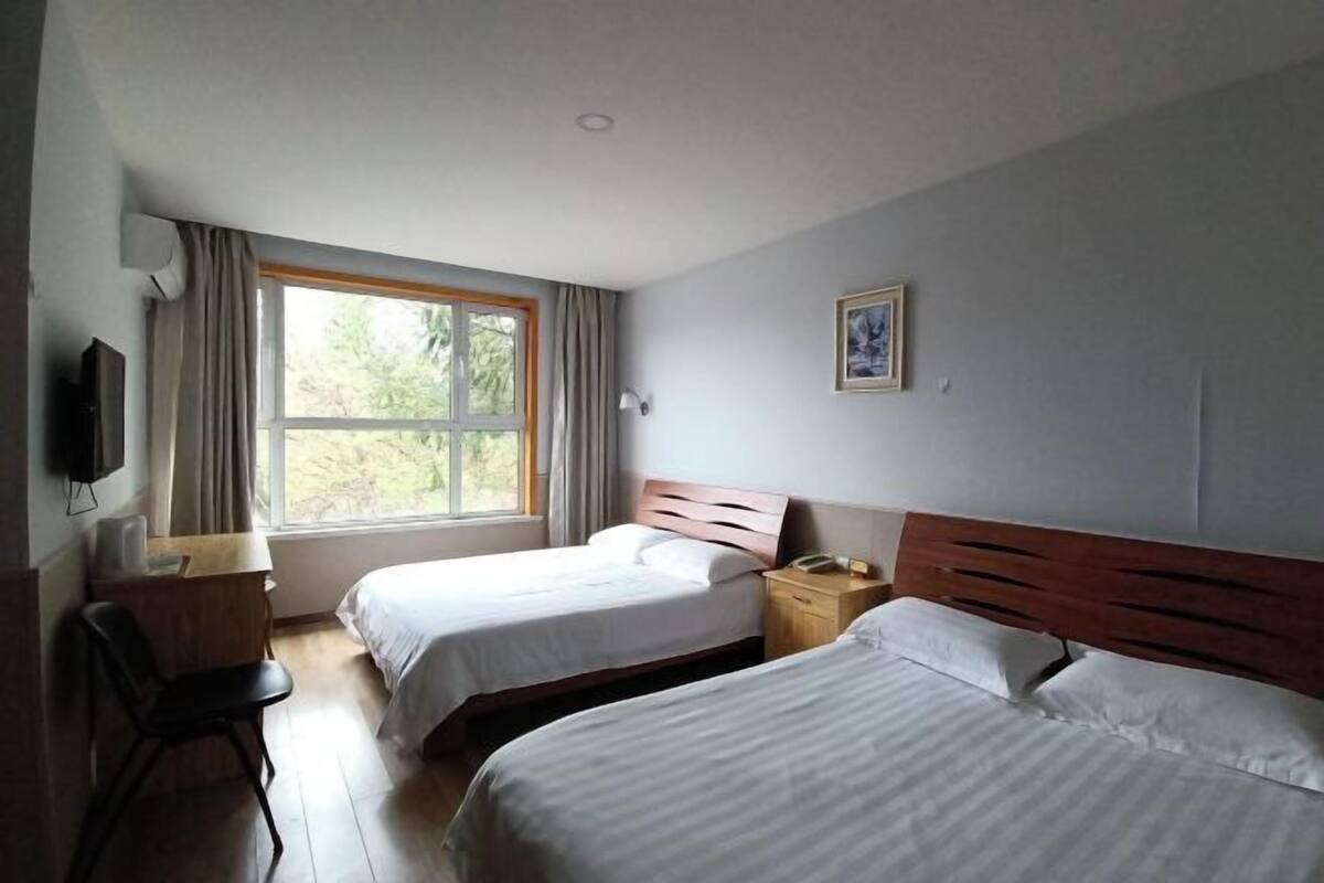 A hotel room with two beds, a desk, a chair, a window with curtains, a wall-mounted TV, and a framed picture.