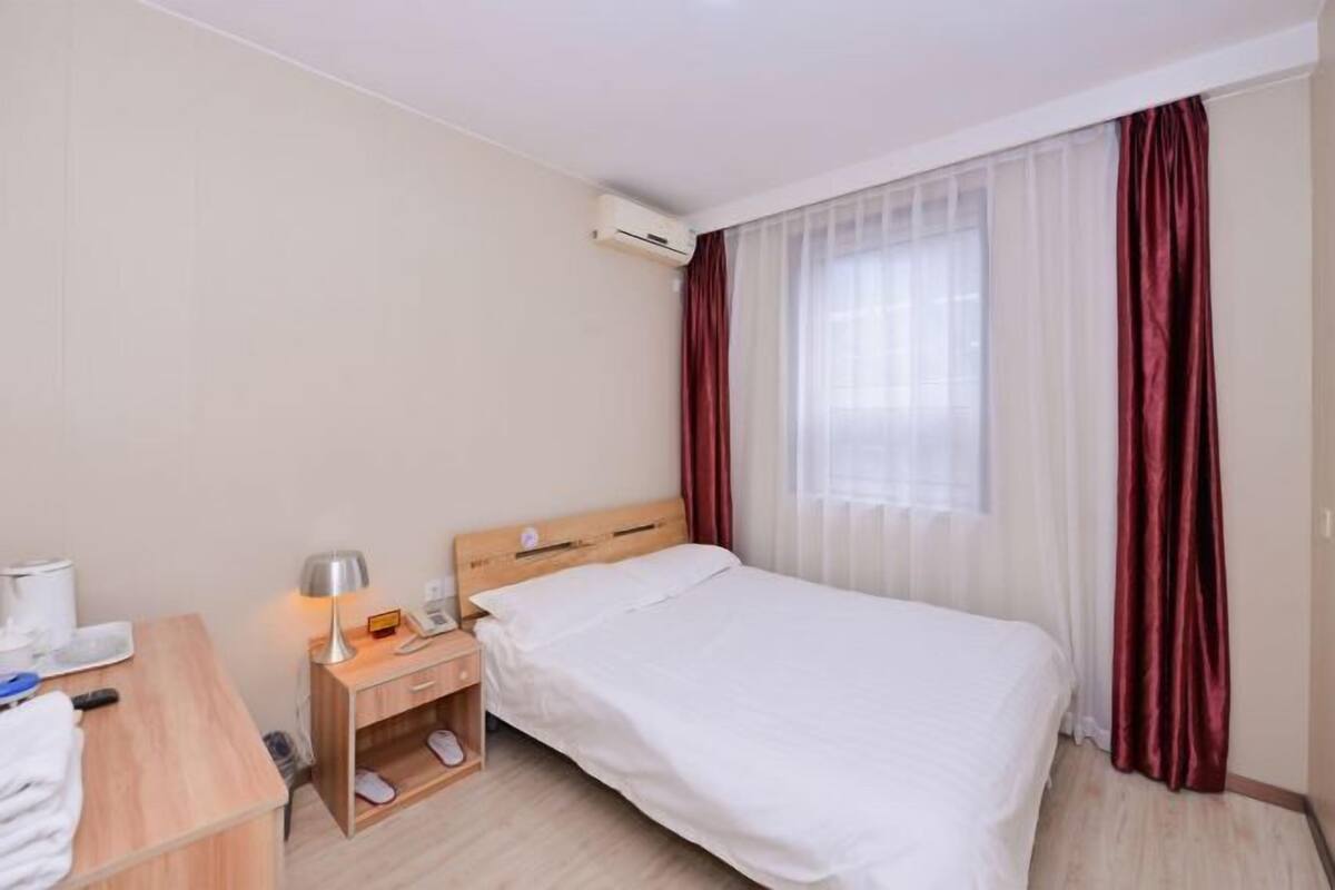 A hotel room with a bed, a bedside table with a lamp, a small shelf, and an air conditioning unit.