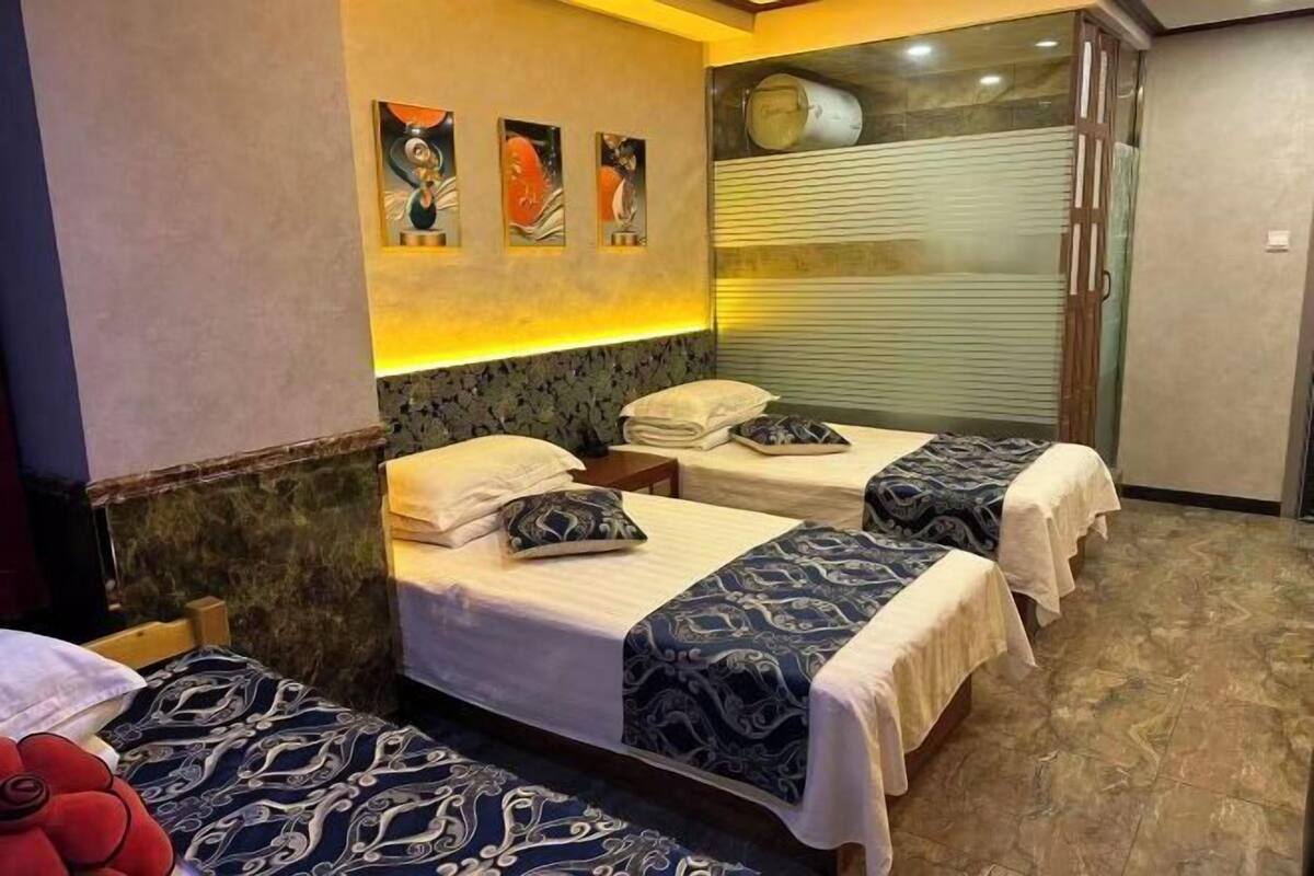 Habitación de hotel con tres camas, cada una con colcha estampada, cabecero y mesita de noche. Se observan cuadros colgados en la pared sobre las camas y un ventanal con persianas.