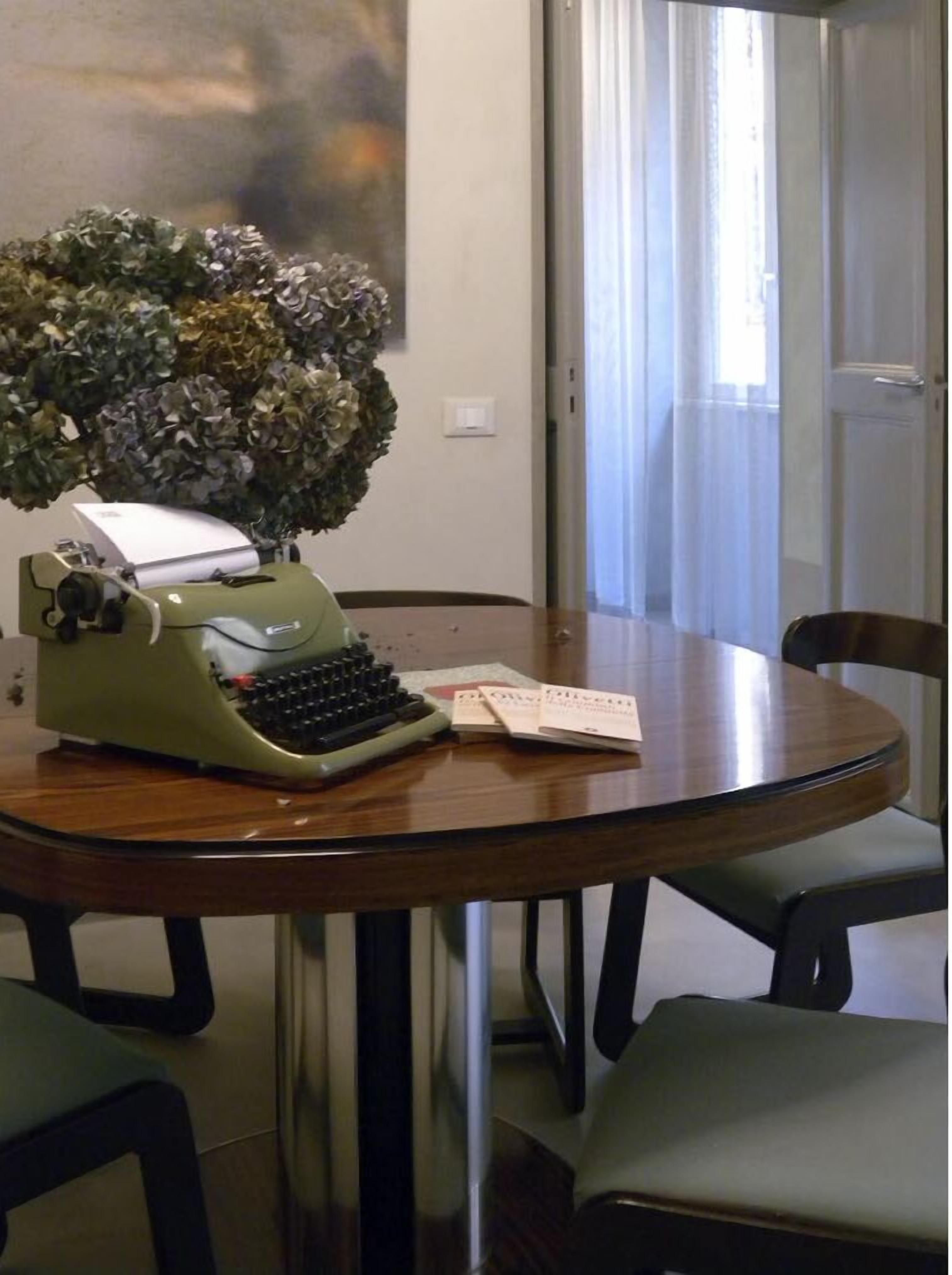Una mesa redonda de madera con una máquina de escribir verde antigua, una planta en maceta y algunos papeles.