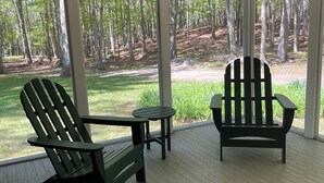 A covered porch with two Adirondack chairs and a small table, overlooking a forested area.