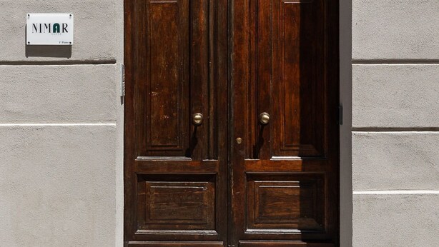 A double wooden door with a metal grille, set in a building with a textured facade. - Nimar suite (Formia)