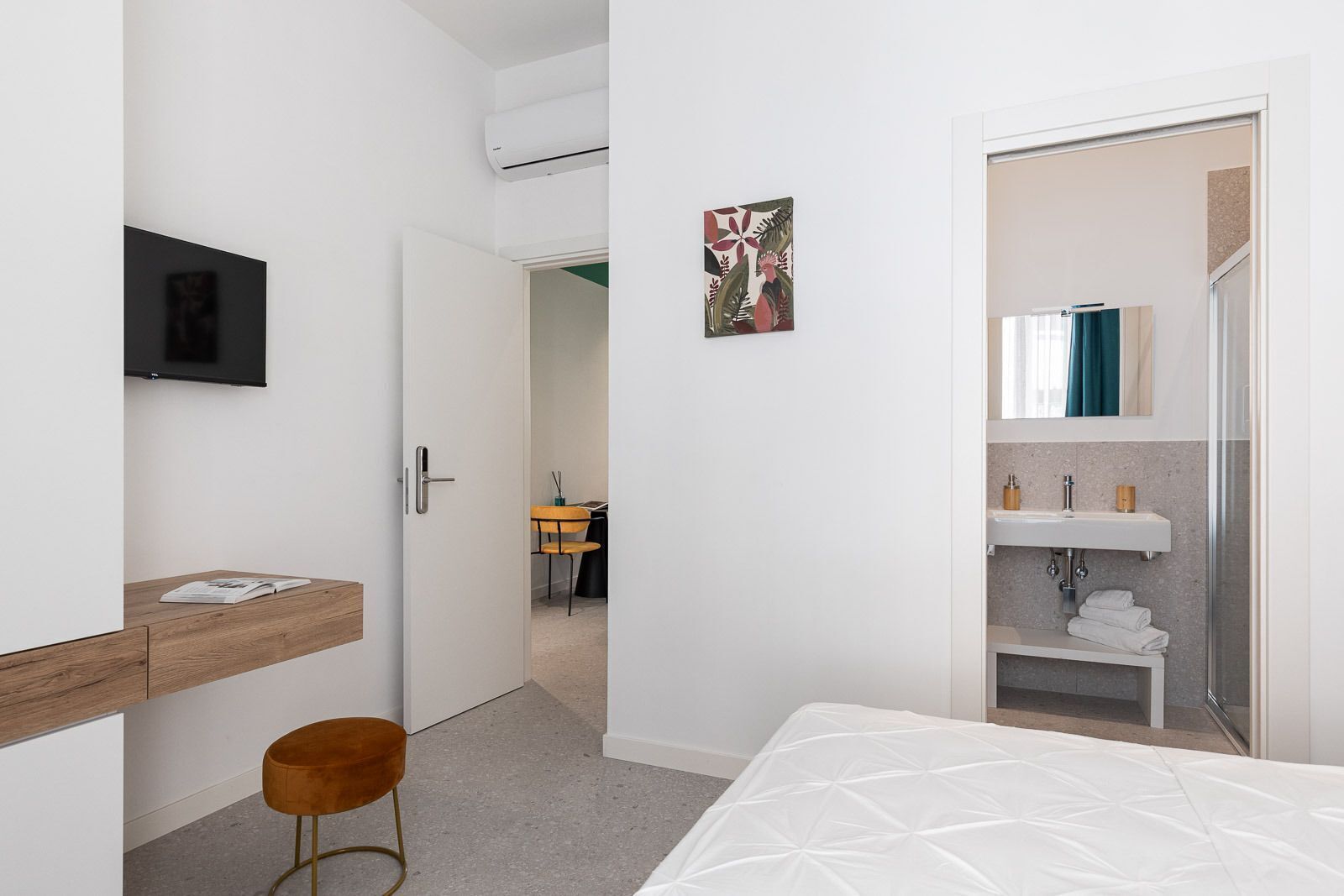 Moderni hotellihuone, jossa on sänky, työpöytä tuolilla, televisio, kylpyhuone, jossa on pesuallas ja peili, sekä ovi, joka johtaa toiseen huoneeseen.