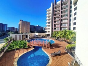 A swimming pool with a wooden deck and lounge chairs, surrounded by greenery and a building with multiple balconies. - New 4 star apartment in prime location (2025), Just a few steps away from Bars, Restaurants, Shoppin... (Pampatar)