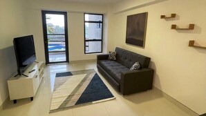 A modern living room with a sofa, a TV on a stand, a large window, and a door leading outside.