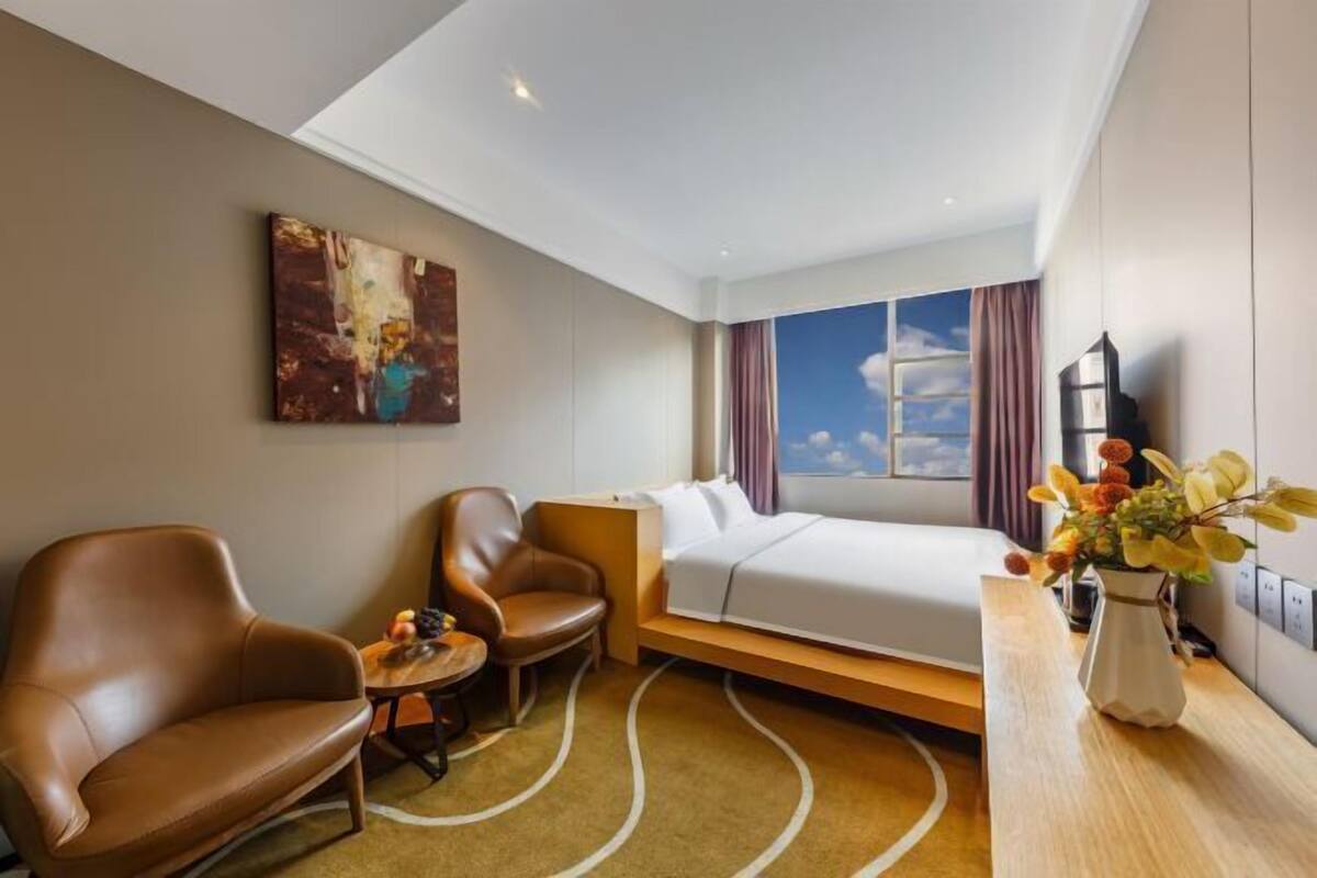 Une chambre d’hôtel avec un lit, deux chaises, une petite table, un tableau et une fenêtre donnant sur le ciel.
