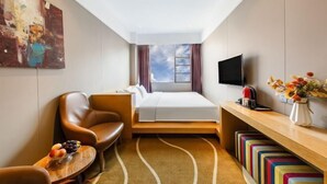 Habitación de hotel con cama, televisor, escritorio con un florero, una silla y un cuadro en la pared.