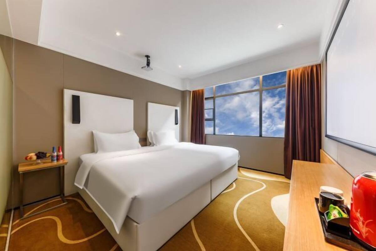 Une chambre d’hôtel avec un grand lit, une table de chevet en bois, une bouilloire rouge et une fenêtre donnant sur le ciel.