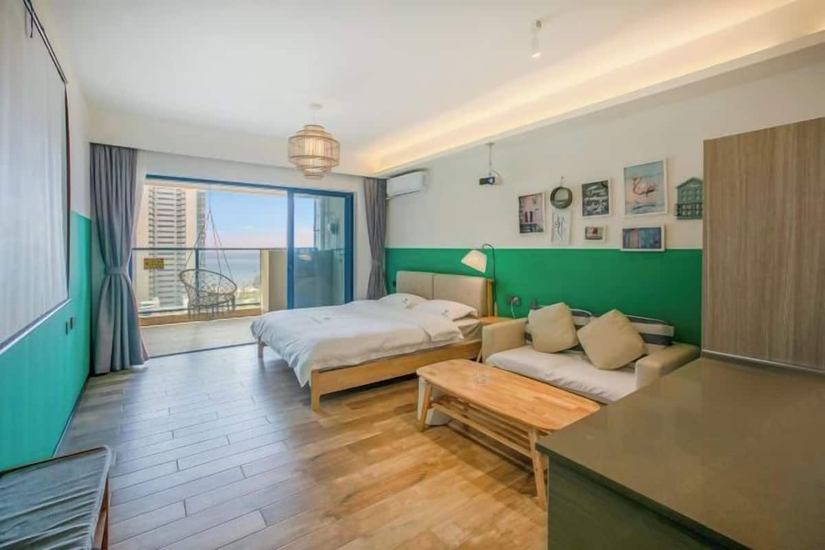 A hotel room with a bed, a sofa, a wooden table, a balcony with a chair, and a city view.
