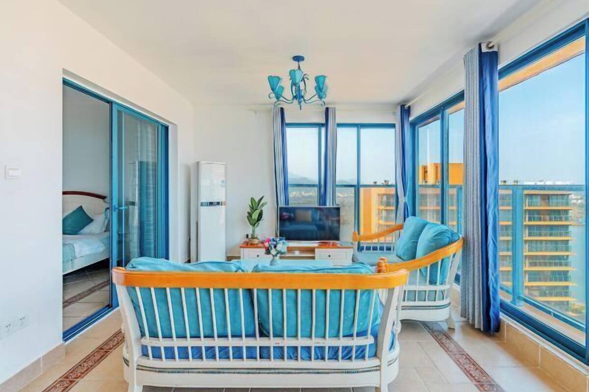 A room with a bed, a sofa, a TV, a chandelier, and large windows.