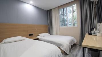 Una habitación de hotel con dos camas individuales, cabecera de madera, ventana con cortinas y escritorio con lámpara y una taza.