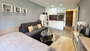 Ein moderner, kompakter Wohnraum mit grauem Sofa, einem Bett, einem kleinen runden Tisch und einer Küchenzeile.