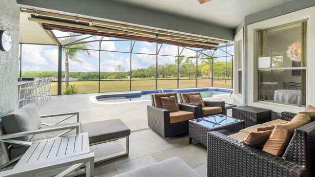 A covered outdoor patio with wicker furniture, a glass table, and a view of a swimming pool and greenery. - EV_149 - 5 Bed 3 Bath Home with Private Screened-In Pool, Games Room (Davenport)