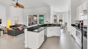 A modern kitchen with white cabinets, a black countertop, and stainless steel appliances. There is a dining area with a table and chairs, and a living room with a sofa and a TV. - EV_149 - 5 Bed 3 Bath Home with Private Screened-In Pool, Games Room (Davenport)