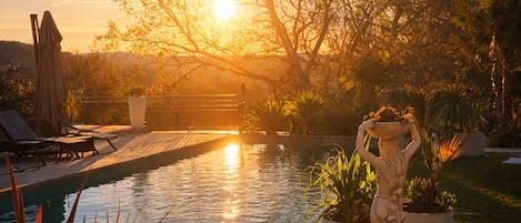 A swimming pool with a statue, surrounded by plants and trees, during sunset.