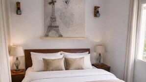 A neatly made bed with a wooden headboard, two bedside tables with lamps, a wall-mounted artwork of the Eiffel Tower, and a window with curtains. - Detached 2-bedroom house with private garden and terrace.Quiet location and pool (Pertuis)