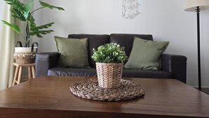Un salón con un sofá marrón oscuro, dos cojines verdes, un camino de fibras naturales con una planta en maceta, un atrapasueños en la pared y una lámpara de pie.