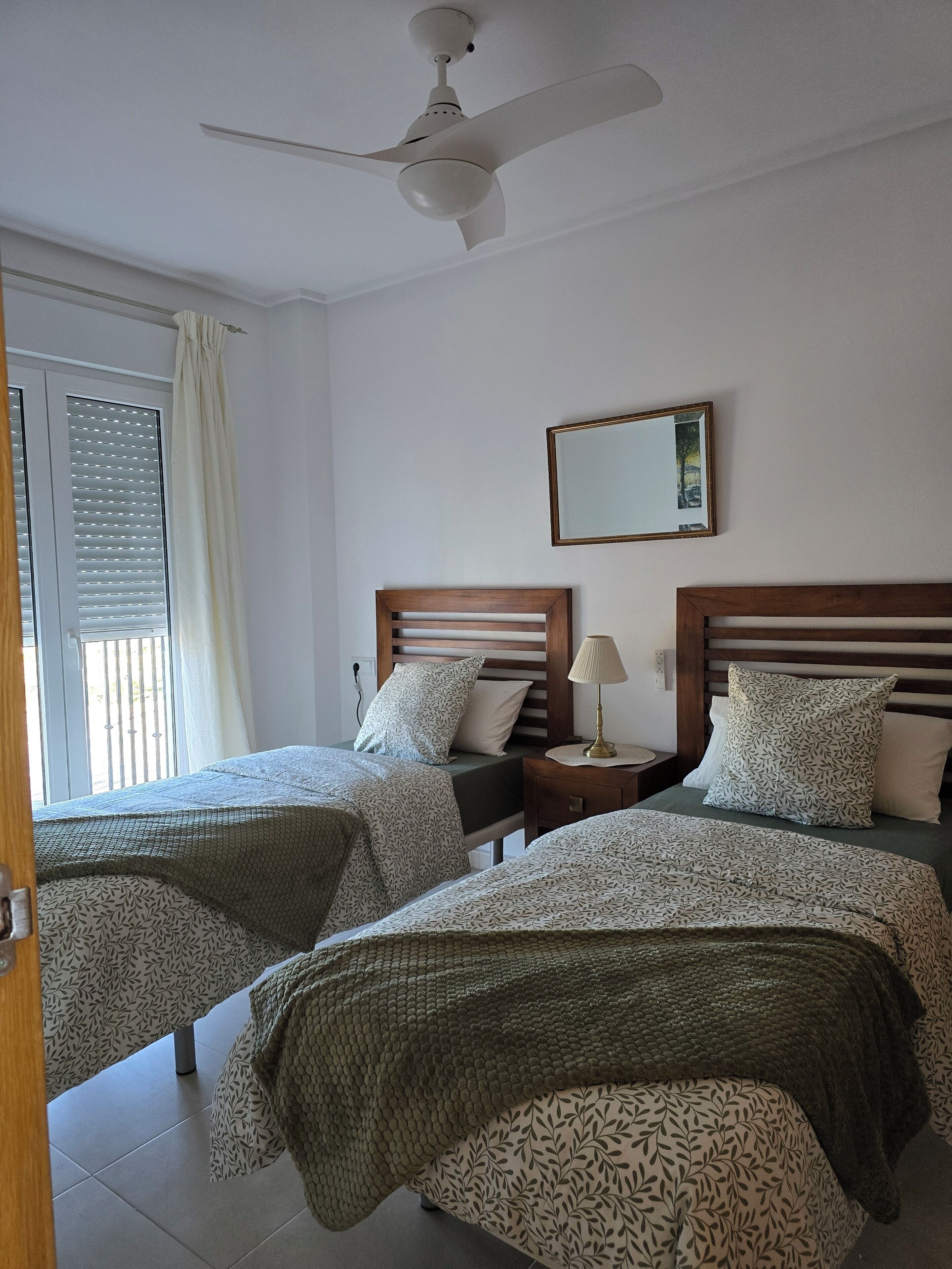 Een hotelkamer met twee bedden, een plafondventilator, een nachtkastje met een lamp, een raam met zonwering en een spiegel aan de muur.