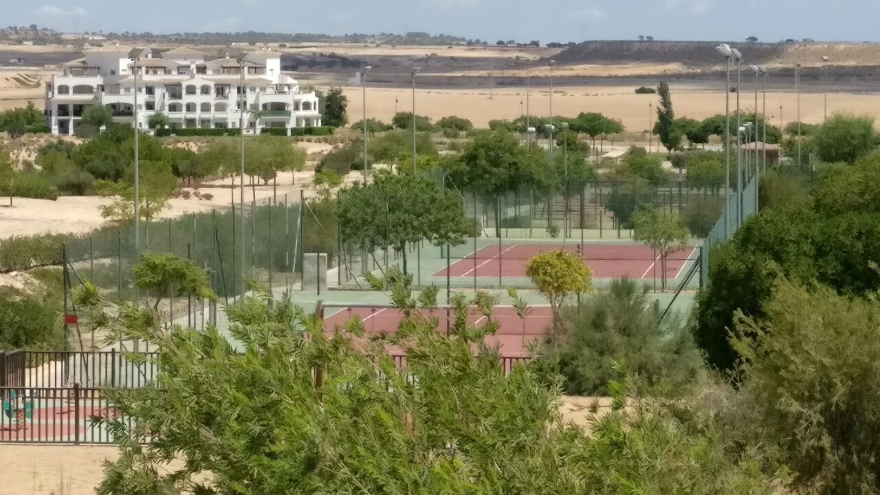 Un court de tennis entouré de verdure, avec un bâtiment en arrière-plan.